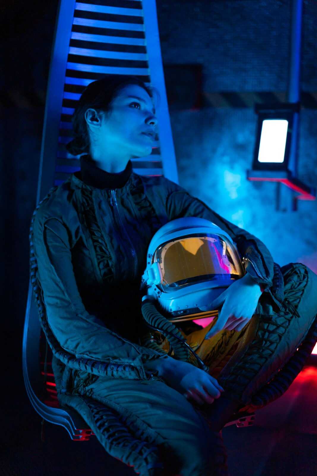 Woman in astronaut suit sitting in a modern, blue-lit space environment.